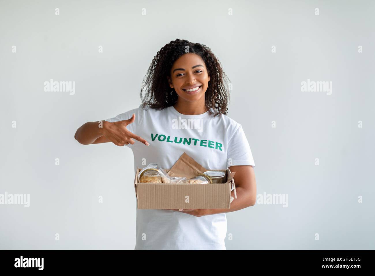 African american female volunteer holding and pointing at box with food donations, posing over light studio background Stock Photo