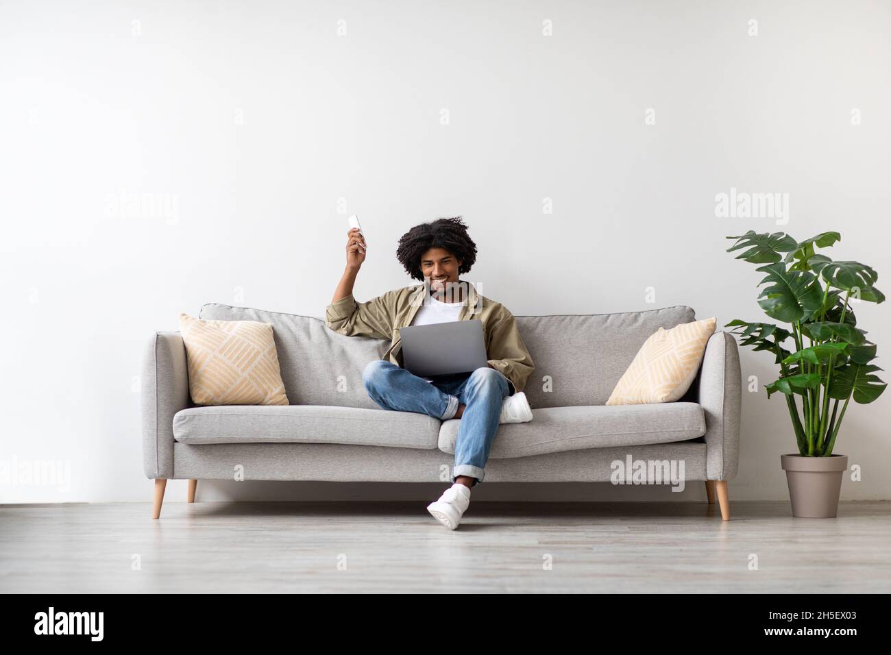 Young Black Man Sitting On Couch With Laptop And Using Remote Controller Stock Photo