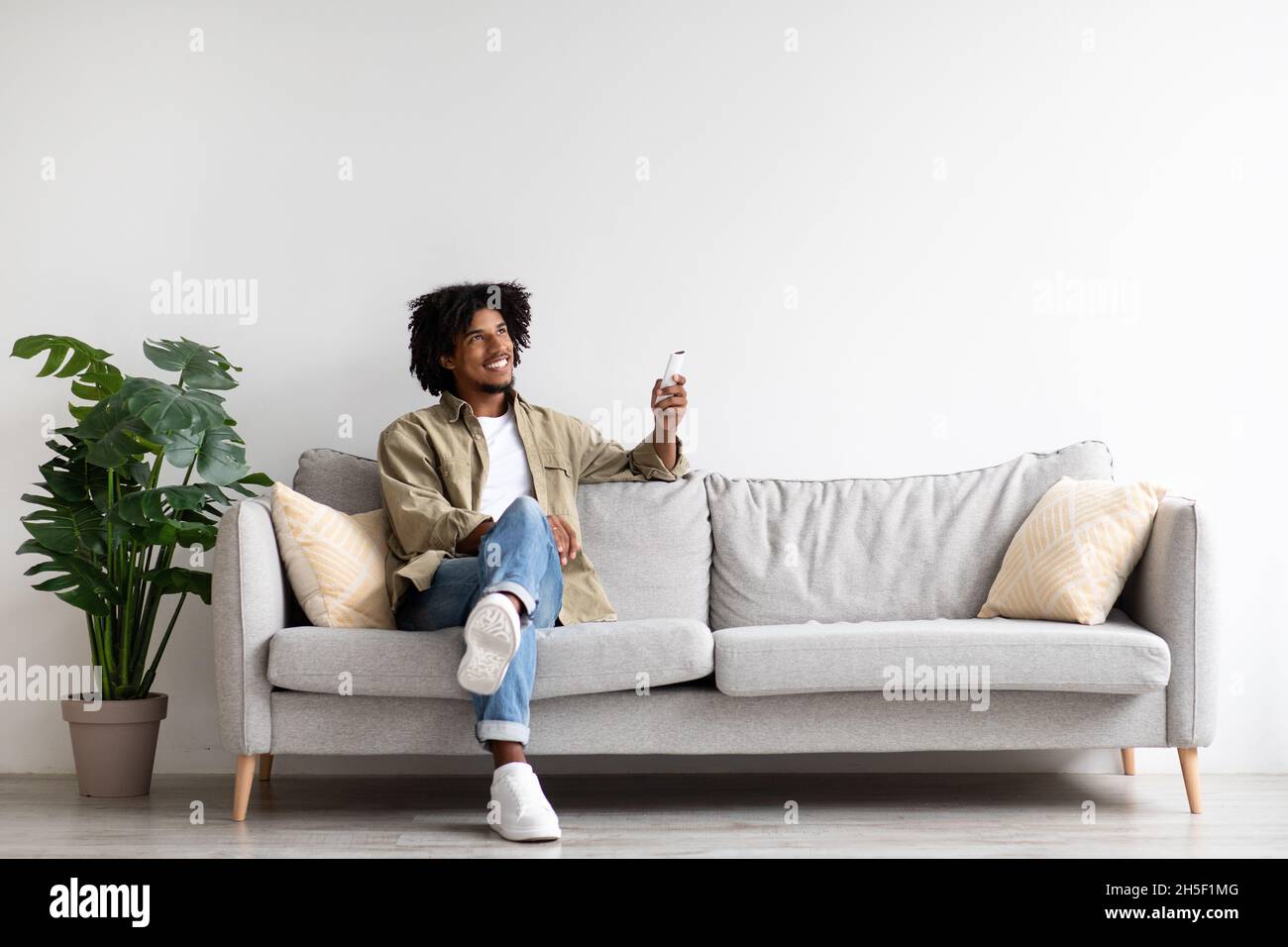 Happy Black Guy Sitting On Couch, Operating Air Conditioner With Remote Controller Stock Photo