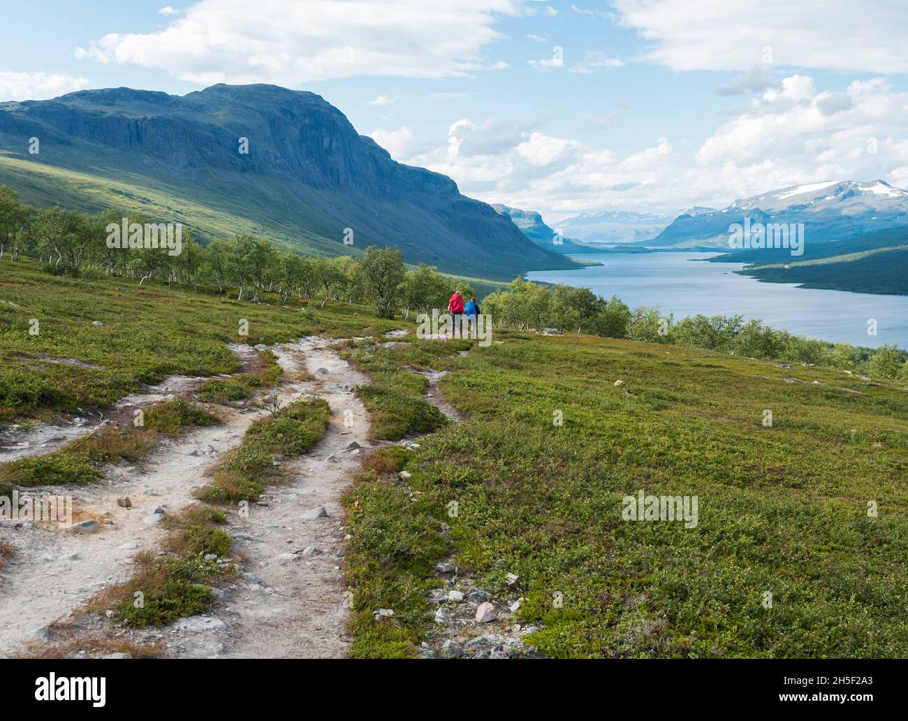 Lapland landscape with beautiful river Lulealven, snow capped mountain, birch tree and footpath of Kungsleden hiking trail with couple of hikers near Stock Photo