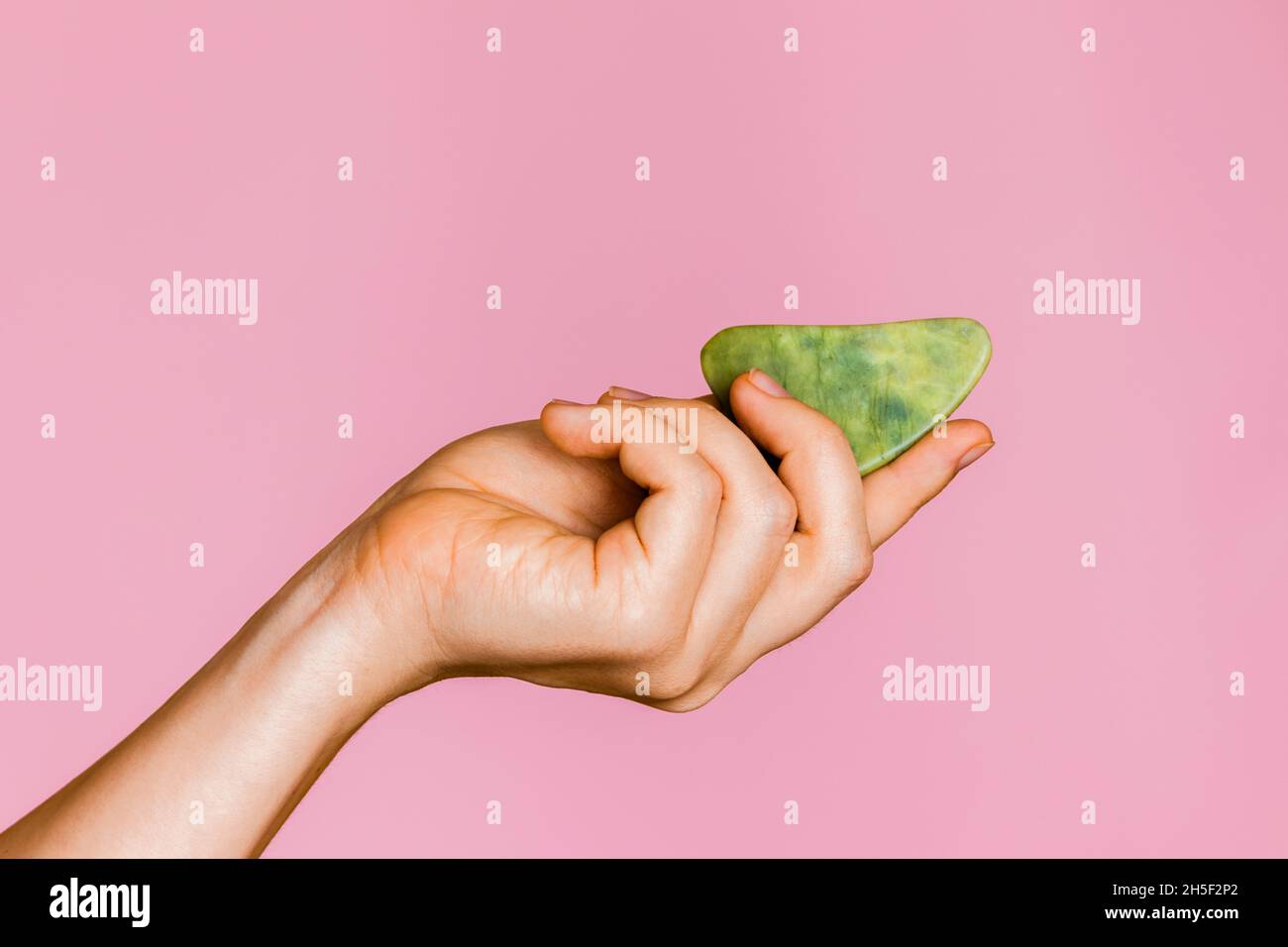 Green jade massager Guasha face scraper in a female hand on a pink background.  Stock Photo