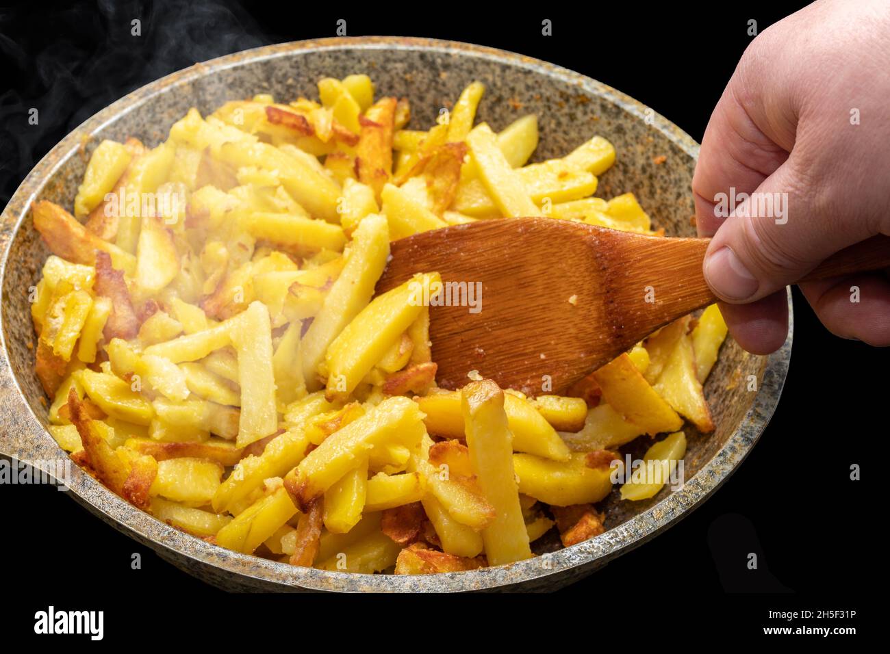 Chef's hand stirring with a wooden spatula delicious crispy golden hot fried potato wedges on an old metal frying pan close-up. Cooking homemade fried Stock Photo