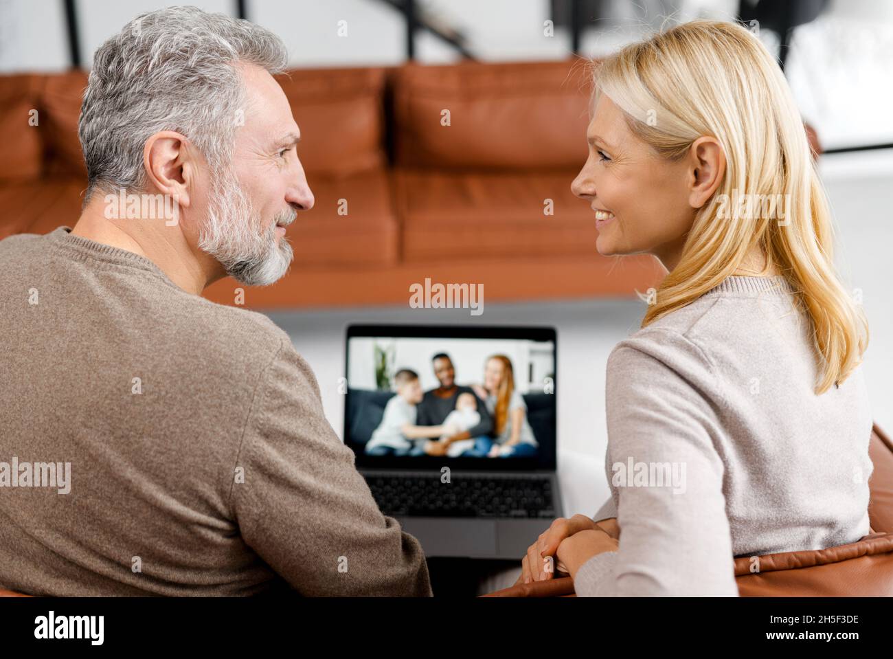 Middle-aged couple using laptop computer for video connection with a grown daughter and her multiracial family, grey-haired man and woman talking online with family on computer screen Stock Photo
