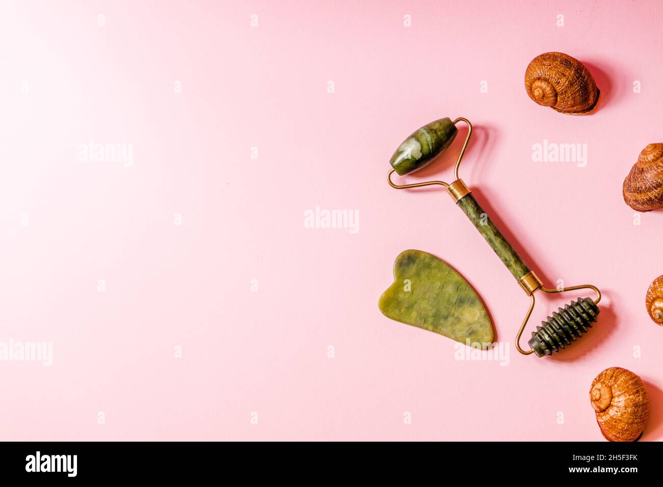 A green jade roller and facial massage scrubber lie on a soft pink background. Stock Photo