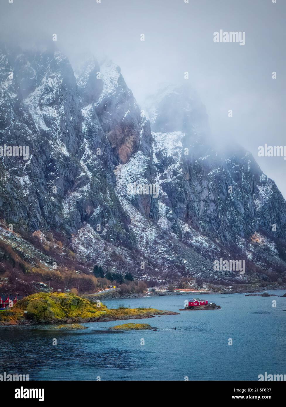 A typical Norwegian landscape scene, with mist-shrouded, snowy granite mountains looming over a tiny red house, isolated in a clear blue fjord. Stock Photo