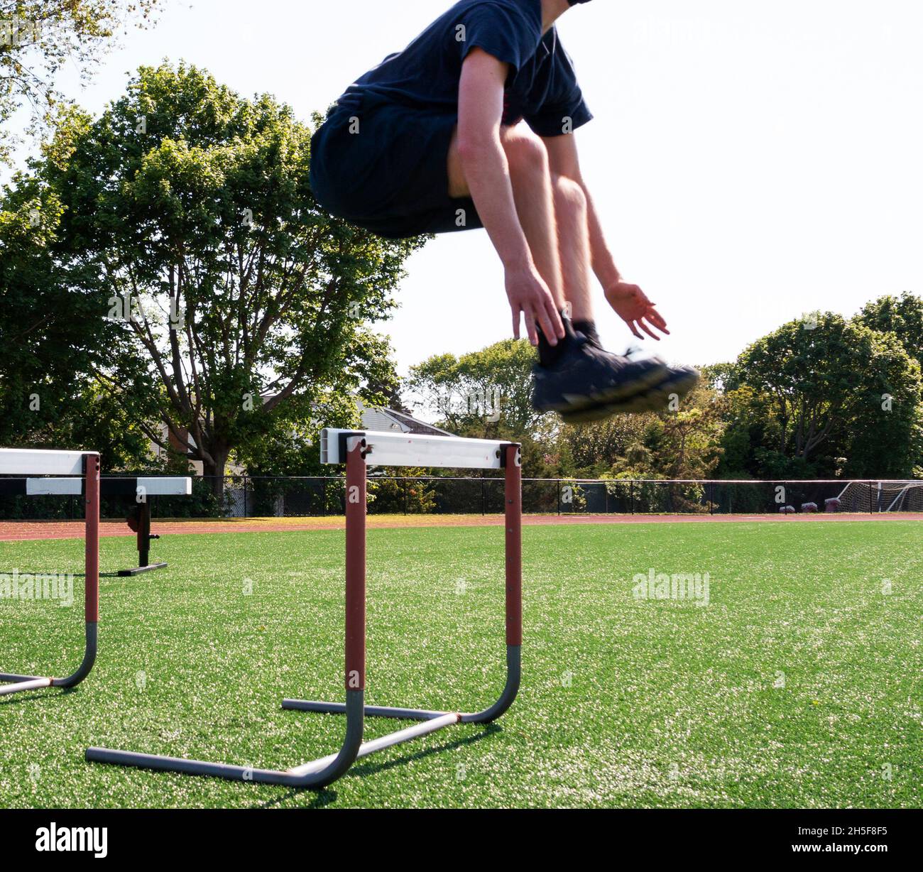 High school athlete jumping over track hurdles on a green turf field at track and field practice. Stock Photo