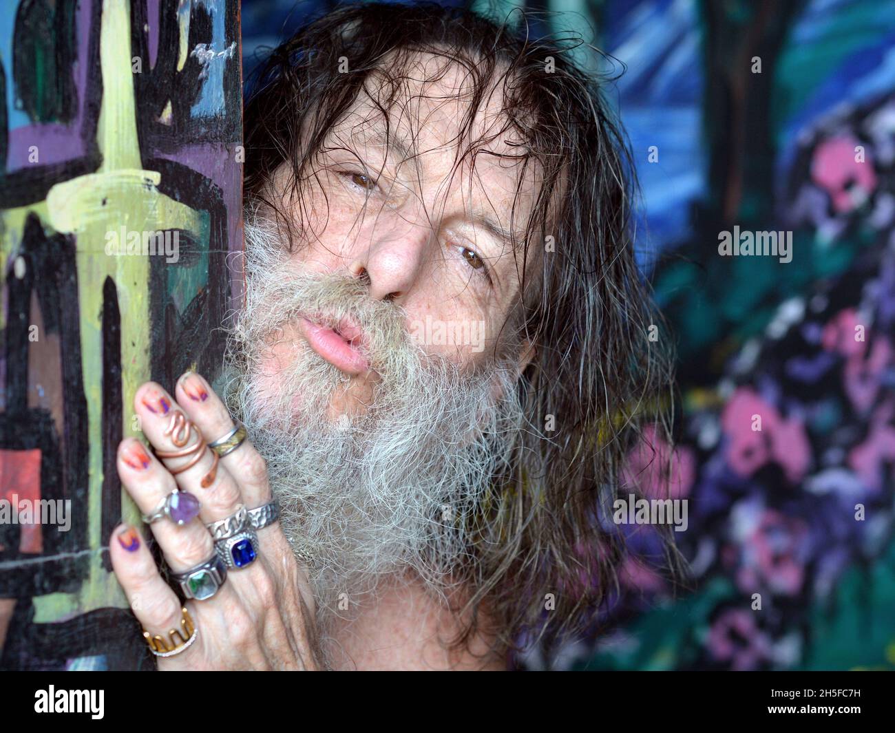 Eccentric old Caucasian man with grey beard, long unkempt hair and multi-ringed hand peeks around a painted door and puckers his lips. Stock Photo