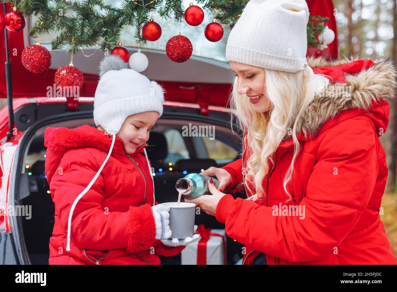 Happy family of blonde mom and little girl sit in christmas decorated car in winter forest and drink hot beverage. Togetherness, holidays, family Stock Photo