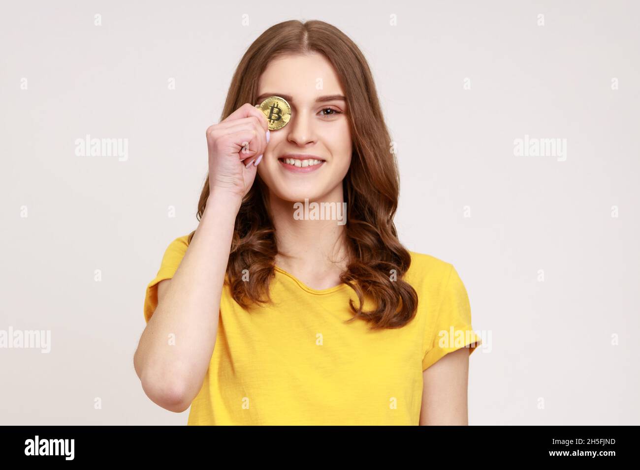 Digital money, cryptocurrency. Attractive woman of young age in casual  style yellow T-shirt, covering eye with golden bitcoin, looking at camera.  Indoor studio shot isolated on gray background Stock Photo - Alamy