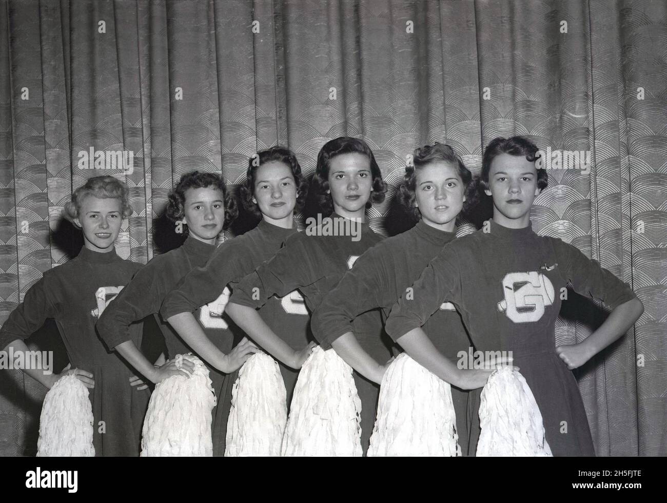 1950s, historical, against a curtain, six american girl cheerleaders in their uniforms and holding their pom-poms, standing hands on hips, in a line for a photo, USA. Cheerleaders have been a part of American team sports, both amateur and professional for decades, particularly in basketball and American football, where girls wear special outfits and wave their pom-poms as they cheer and support their teams. In this era, the first letter of the high school the cheerleaders attended would often be embroidered onto the outfits - as we see here the letter G - in the middle of a megaphone shape. Stock Photo