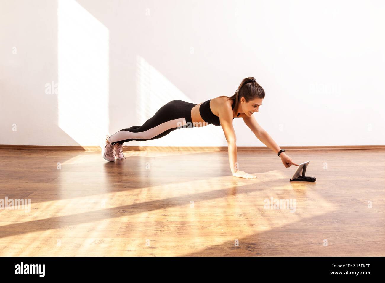 Slim woman doing plank exercise, repeat after coach online, choosing next tutorial video on tablet, wearing black sports top and tights. Full length studio shot illuminated by sunlight from window. Stock Photo