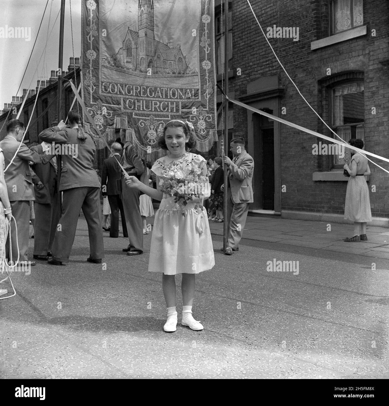 1950s, historical, outside in a street of victorian terraced houses, a young girl standing for her photo holding a rippon attached to a pole supporting a large hand-sown decorated blanket for the Congregational Church of Audley Range, Blackburn, Lancashire, England, UK. From the 1840s, the industrial revolution and the growth of the cotton and textile industries had transformed the area and terraced housing, schools, shops, pubs and churches were built on farm land to service the new population working in the factories and textile mills. Stock Photo