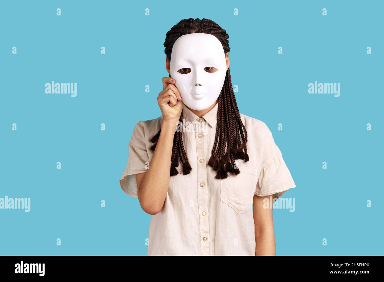 Portrait of anonymous woman with black dreadlocks hiding her face behind white mist, wants to hide his personality, wearing white shirt. Indoor studio shot isolated on blue background. Stock Photo