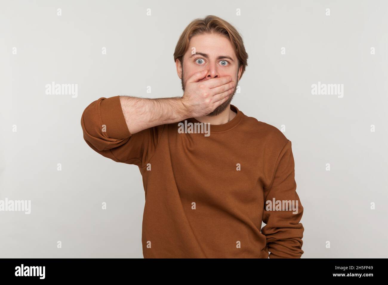 Portrait of scared man with beard wearing sweatshirt, covering mouth with hand, looking with frightened shocked eyes, keeping secret taboo. Indoor studio shot isolated on gray background. Stock Photo