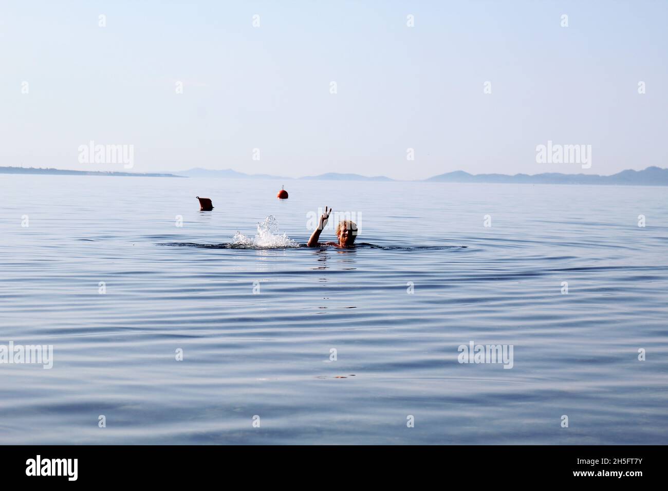 Mann schwimmt im Meer. Kopf eines Mannes, der aus dem Wasser ragt. Der Mann macht ein Peace Zeichen. Bojen im Hintergrund. Vir, Kroatien. Stock Photo
