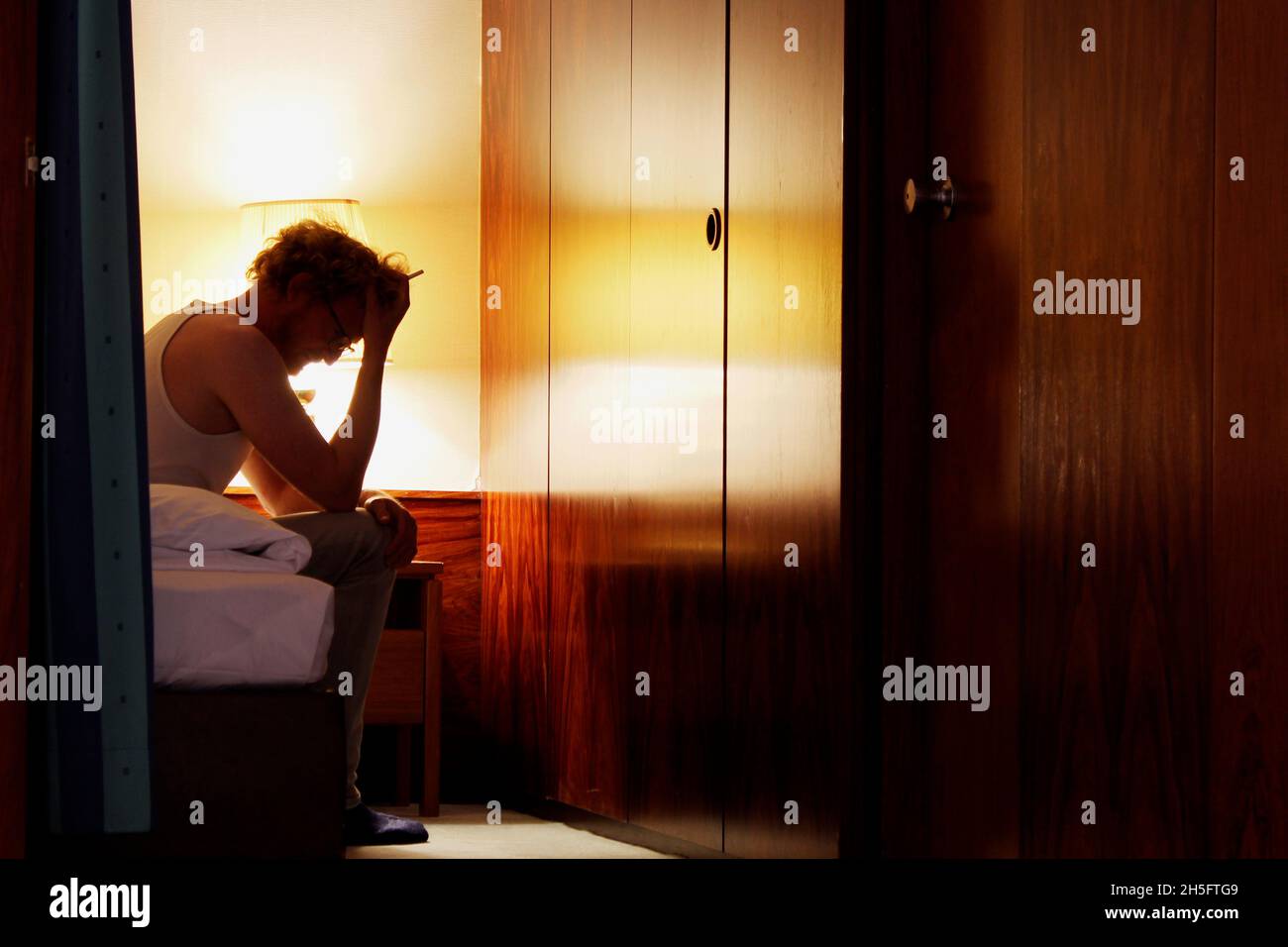 Ein Mann in einem weißen Unterhemd sitzt alleine im Hotelzimmer, den Kopf gesenkt, beide Hände vor dem Gesicht, mit einer Zigarette in der Hand. Stock Photo