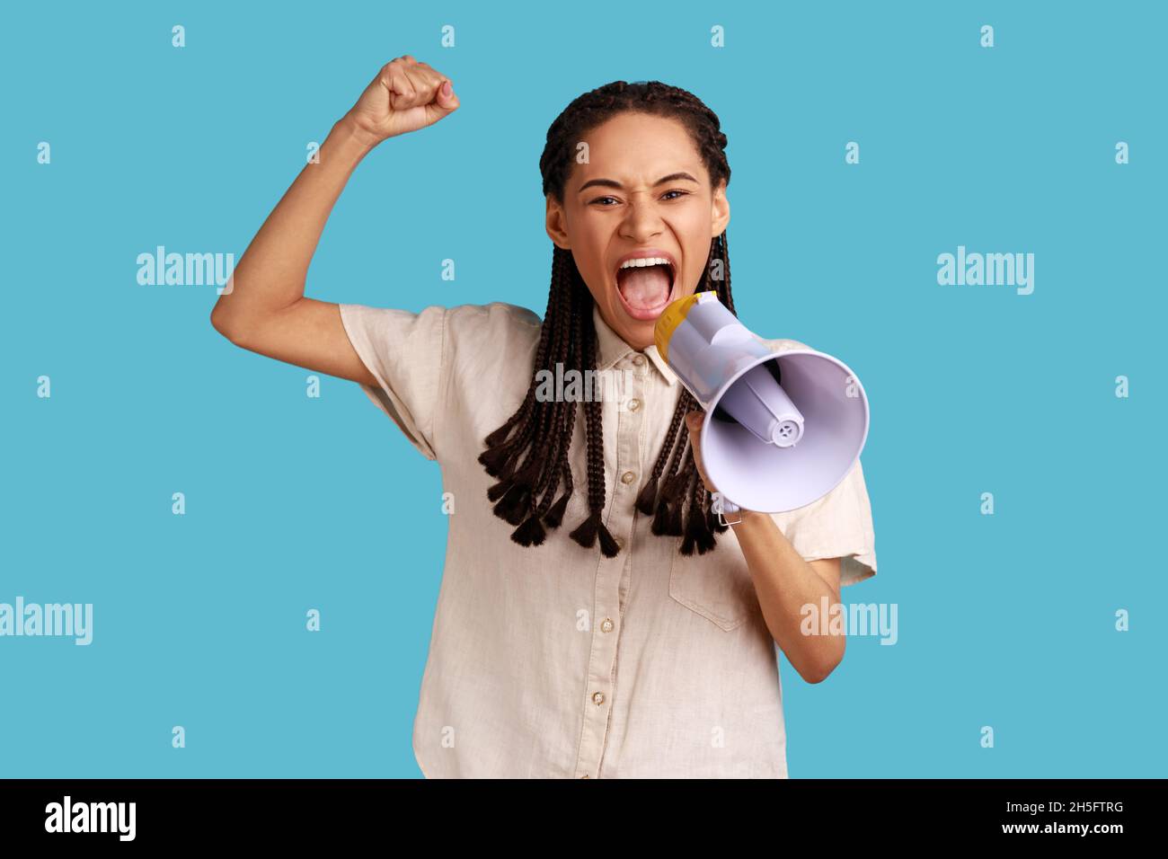 Portrait of woman with black dreadlocks standing with raised hands and holding megaphone, screaming in loud speaker, protesting, wearing white shirt. Indoor studio shot isolated on blue background. Stock Photo