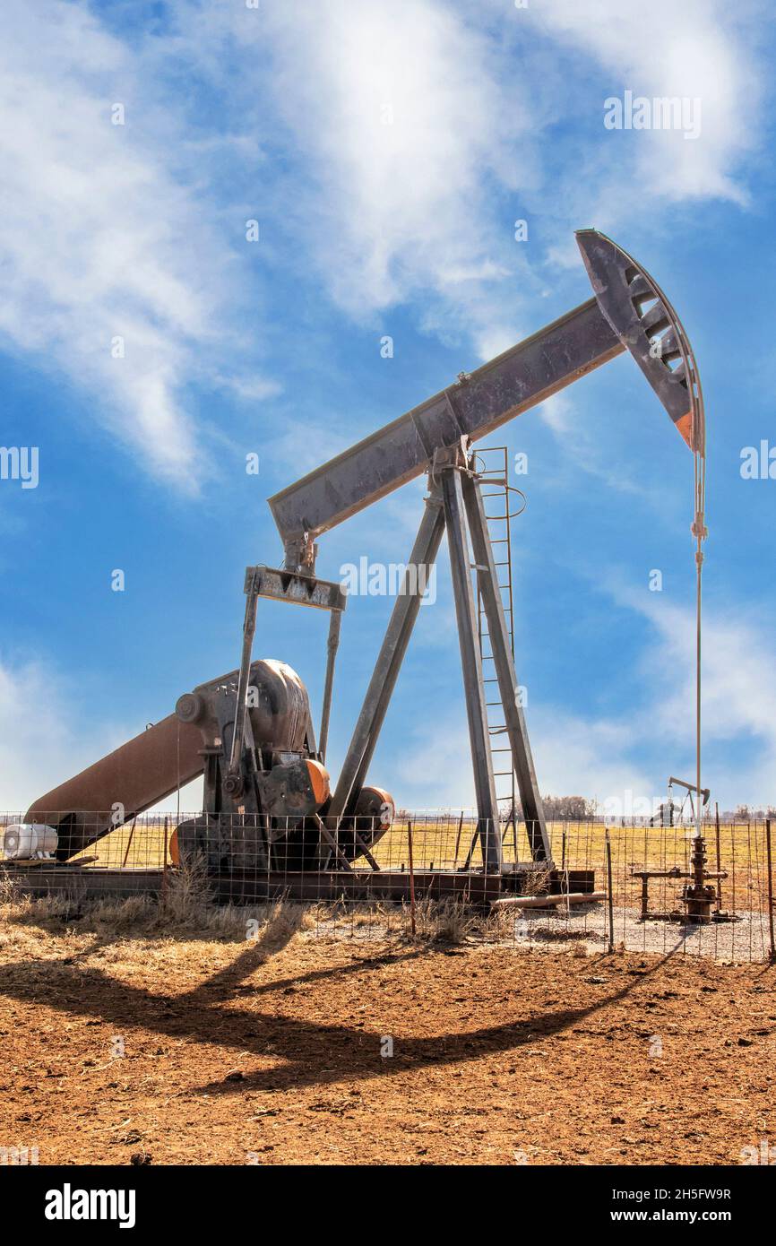 Oilfield pump jack out in summer red dirt field on bright day with shadow and a few whispy clouds in blue sky. Stock Photo