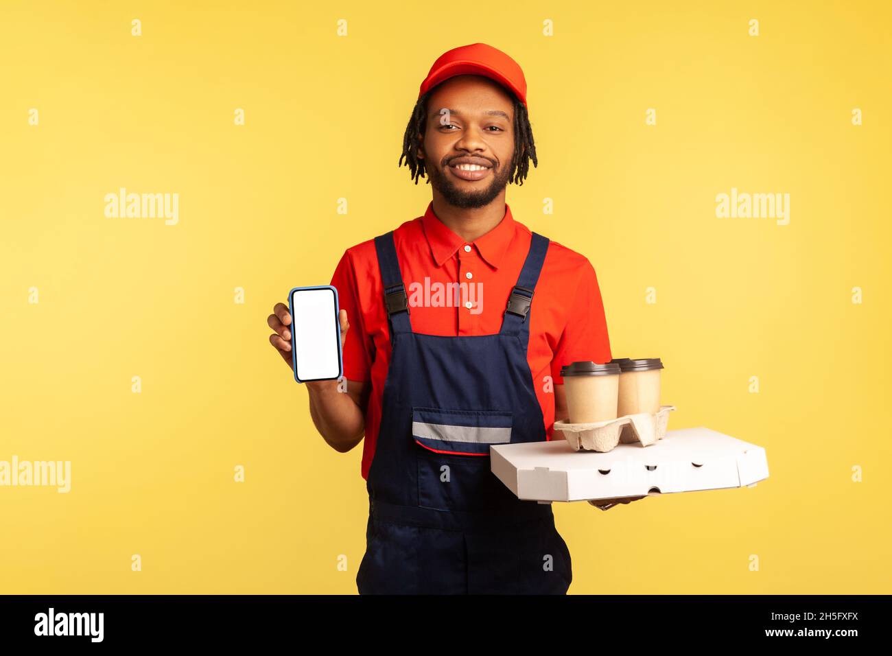 Delivery mobile app, internet advertise. Friendly courier man in overalls holding coffee cups and cellphone with mock up display, online order service. Indoor studio shot isolated on yellow background Stock Photo