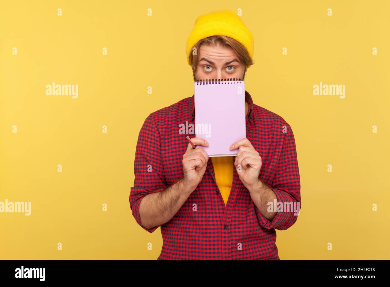 Portrait of hipster bearded guy in beanie hat and checkered shirt standing covering half of face with paper notebook, looking at camera. Indoor studio shot isolated on yellow background. Stock Photo