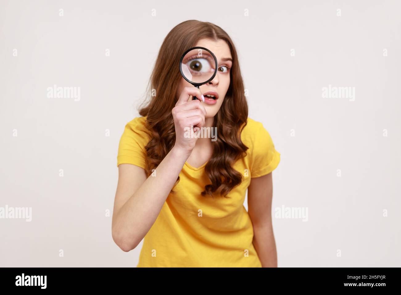 Portrait of funny young female in yellow casual T-shirt holding magnifying glass and looking at camera with big zoom eye, curious face. Indoor studio shot isolated on gray background. Stock Photo