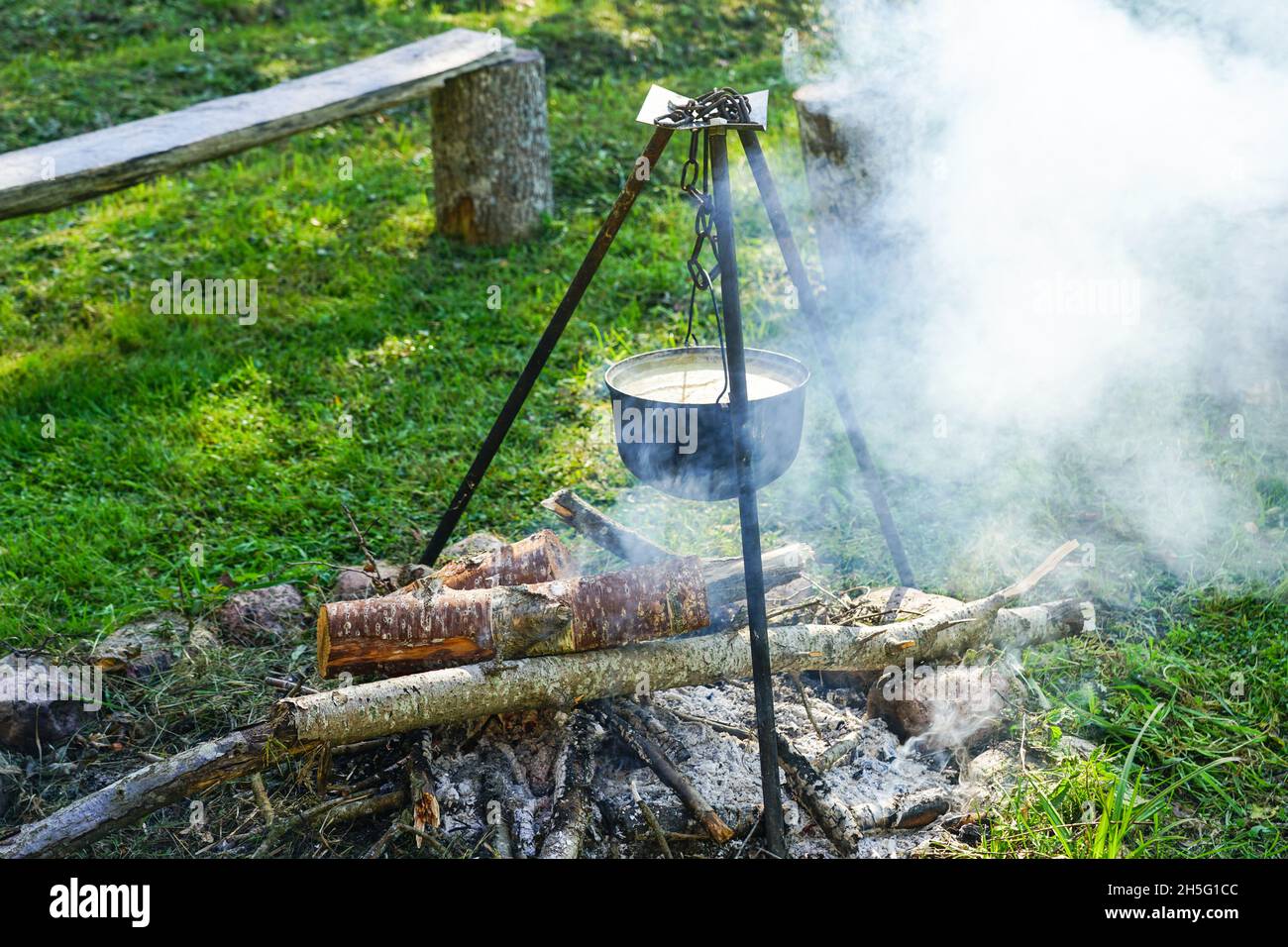 https://c8.alamy.com/comp/2H5G1CC/soup-pot-hanging-on-an-iron-tripod-on-a-campfire-in-a-green-meadow-smoke-from-firewood-2H5G1CC.jpg