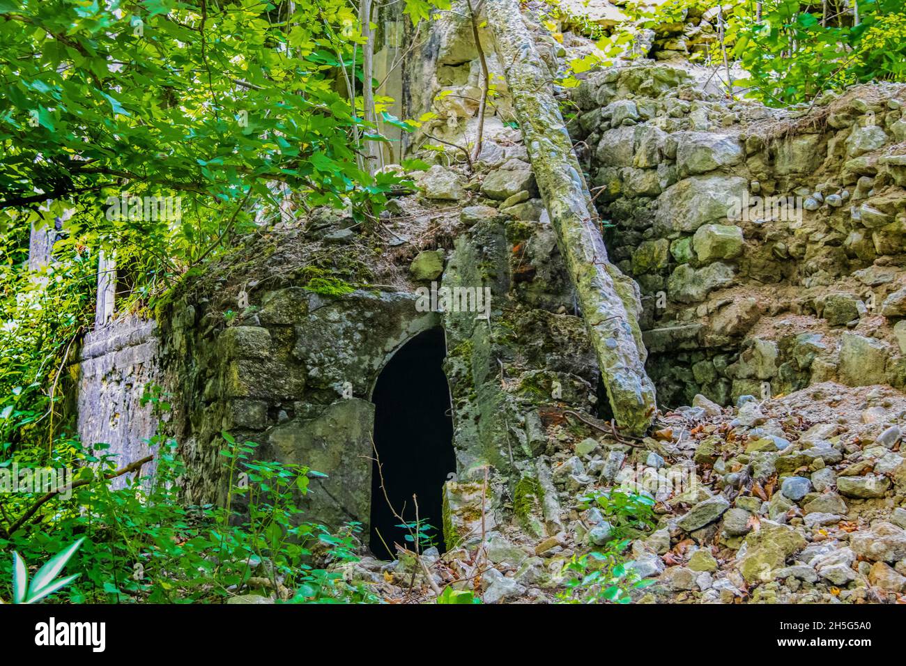 Collapsed rock and concrete bridge with arched opening underneath partly covered and tumbled rocks and debrie and overgrown vegetation Stock Photo