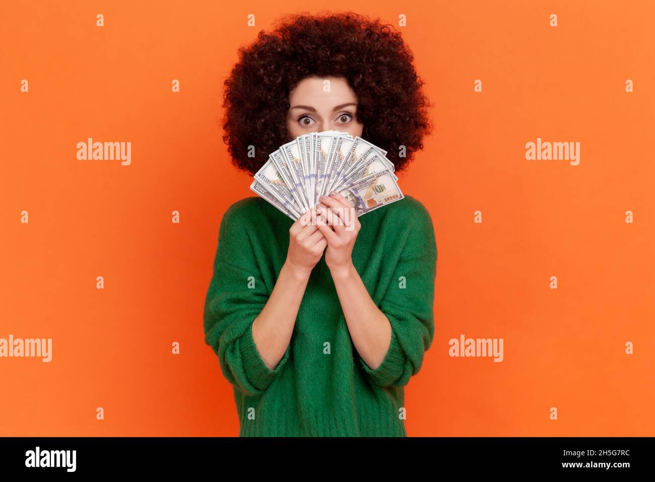 Portrait of woman with Afro hairstyle wearing green casual style sweater covering face with dollars banknotes, hiding, holding currency. Indoor studio shot isolated on orange background. Stock Photo