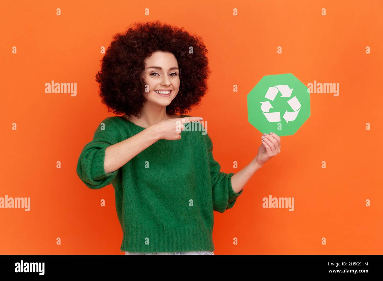 Satisfied woman with Afro hairstyle wearing green casual style sweater pointing at green recycling symbol in her hands, ecology concept. Indoor studio shot isolated on orange background. Stock Photo