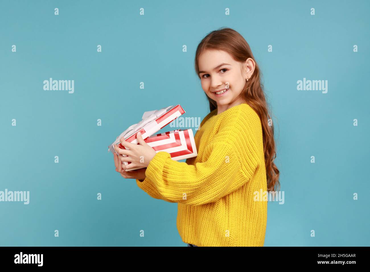 Side view of little girl opens gift box, smiling to camera, celebrating birthday, Christmas holiday, wearing yellow casual style sweater. Indoor studio shot isolated on blue background. Stock Photo