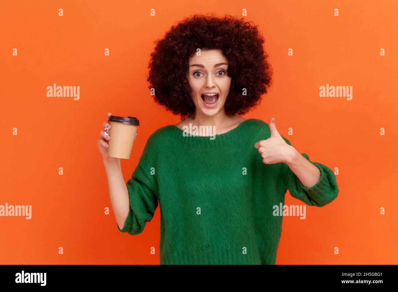 Excited woman with Afro hairstyle wearing green casual style sweater holding coffee to go and showing thumb up, recommend coffee house. Indoor studio shot isolated on orange background. Stock Photo