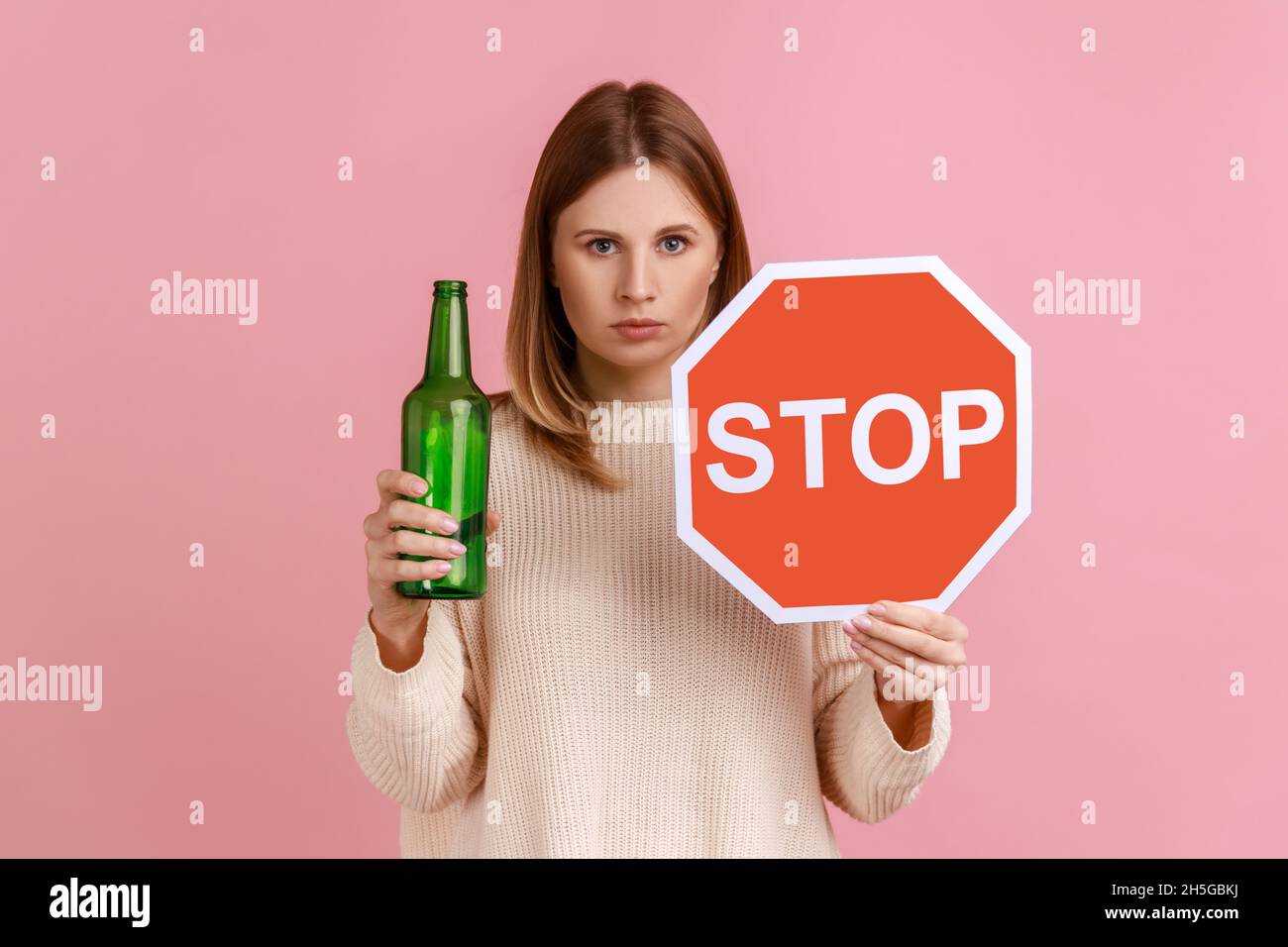 Portrait of serious blond woman holding red stop sign and bottle with alcoholic beverage, calls on dont drink alcohol, wearing white sweater. Indoor studio shot isolated on pink background. Stock Photo