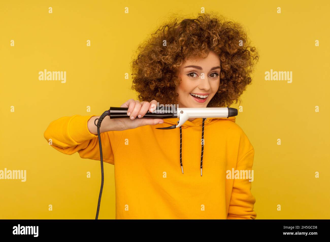 Woman holding curling iron, making perfect afro hairdo, ironing hair curls, grooming with professional tool, wearing casual style hoodie. Indoor studio shot isolated on yellow background. Stock Photo