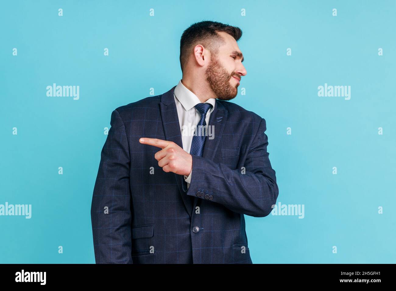 Get out. Portrait of angry man with beard wearing dark suit looking aside and pointing finger another way, asking to leave, dissatisfied expression. Indoor studio shot isolated on blue background. Stock Photo