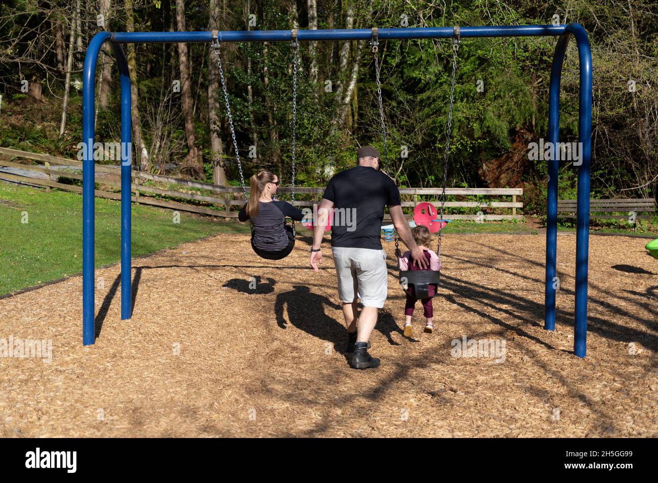 A young family plays at a playground together while the mom and daughter are at the swingset and the dad pushes his daughter on the swing Stock Photo
