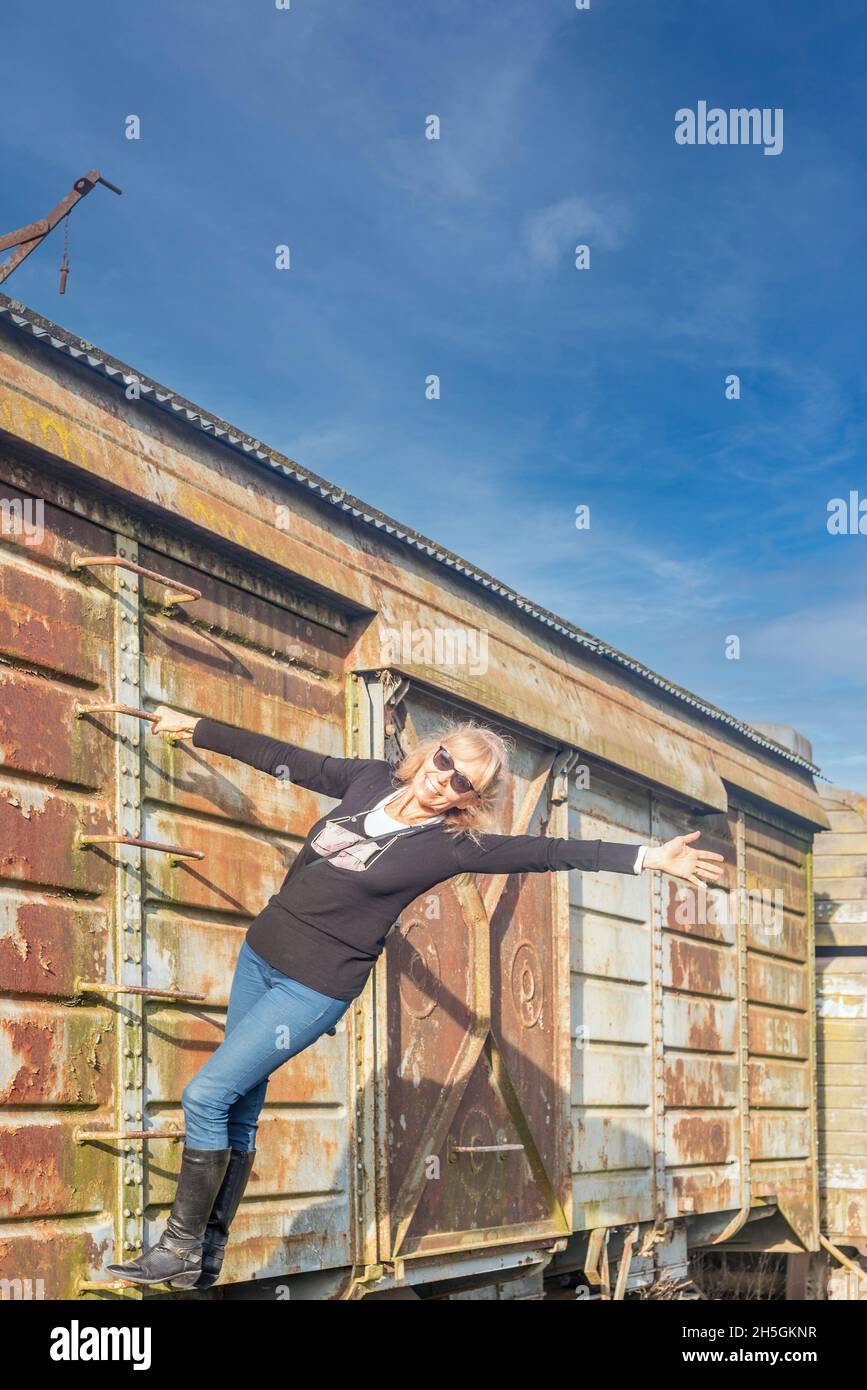 Portrait of a blonde adult woman with sunglasses stand on an abandoned train car ladder smiling and with open arms. Stock Photo