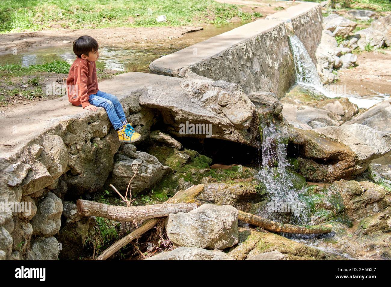 latino brunette child pensive sitting on a ravine of waterfall of a creek, wearing jeans and yellow boots. Boy gazing at the relaxing stream. Horizont Stock Photo