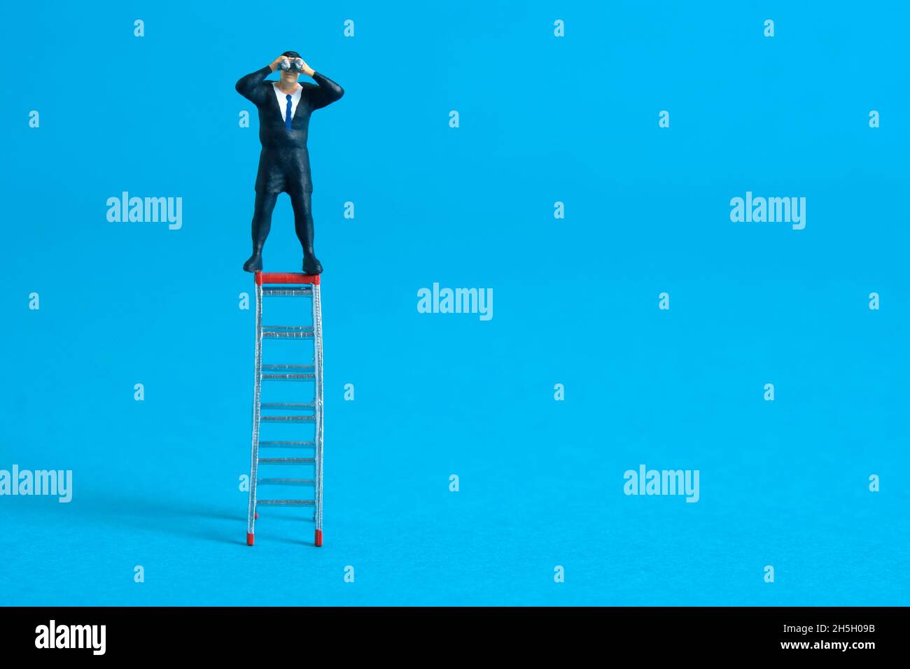 Businessman using binoculars standing above folding ladder. Miniature tiny people toys photography. isolated on blue background. Stock Photo