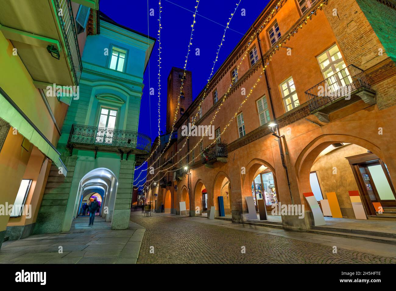 Narrow cobblestone street among historic buildings illuminated by Christmas lights in the evening in old town of Alba, Piedmont, Northern Italy. Stock Photo