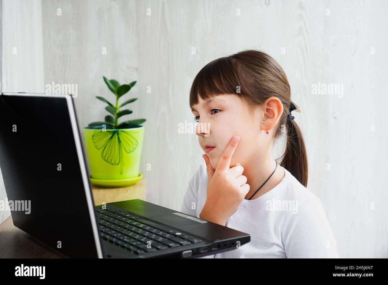 Indoor shot of cute dark haired female child looking at notebook thoughtfully with hand on face Stock Photo