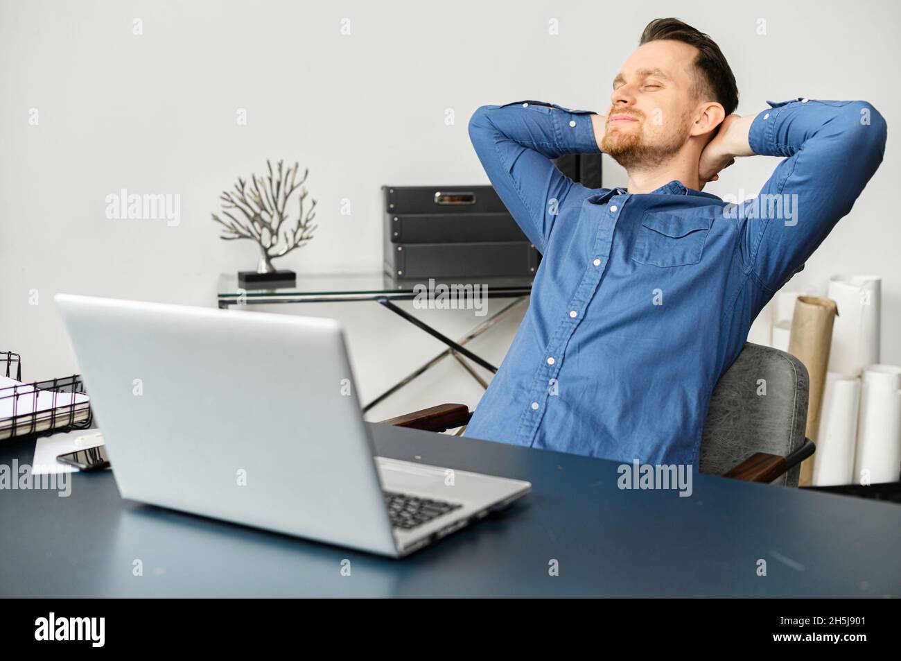 Peaceful millennial guy sitting at the desk in the modern home office, feeling excited for completing the project, placed hands behind head and stretching, enjoying remote freelance job Stock Photo