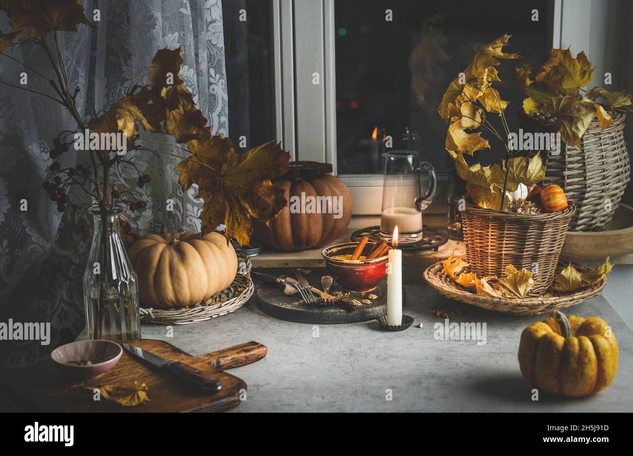 Cozy autumn kitchen background with pumpkins, autumn leaves, candles, cutting board and kitchen utensils on grey concrete table at window. Domestic st Stock Photo