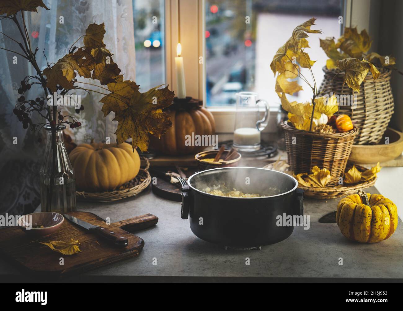 Cooking pot with steam and millet on kitchen table with pumpkins, autumn leaves, candles and kitchen utensils at window background. Cooking healthy gr Stock Photo