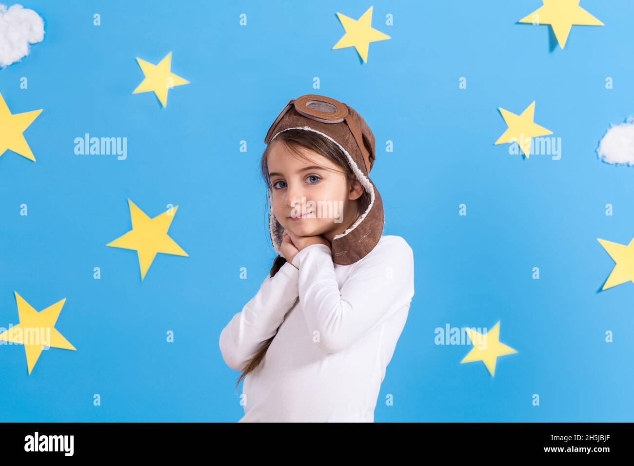 Little happy girl in aviator's cap smiling and playing, in white shirt at the studio. Blue sky, white clouds and stars background. Stock Photo