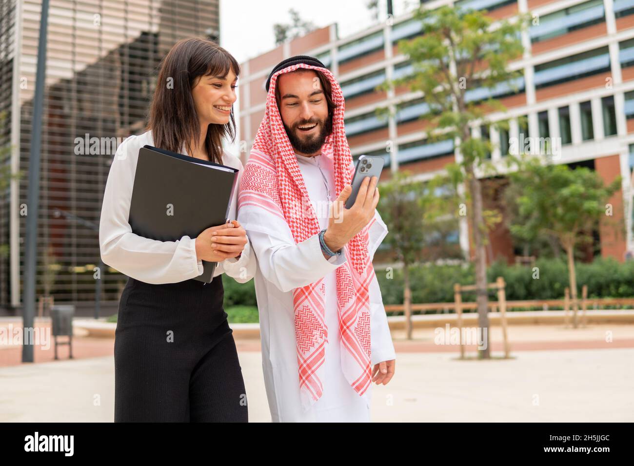 Happy Arab man smiling and showing video on smartphone to businesswoman while standing on street in city downtown Stock Photo