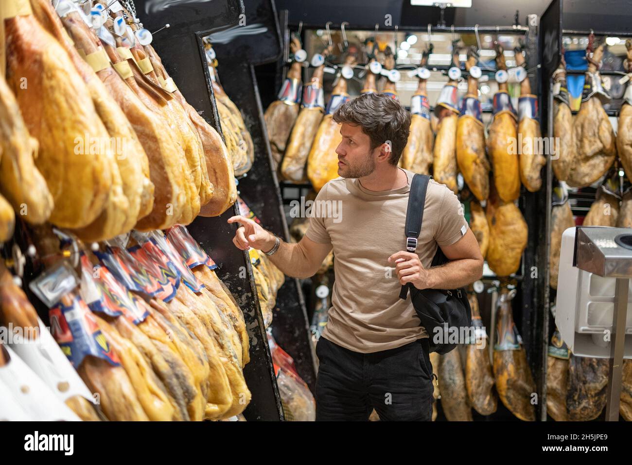 Male client with bag examining and selecting Spanish jamon hanging on racks while shopping in supermarket Stock Photo
