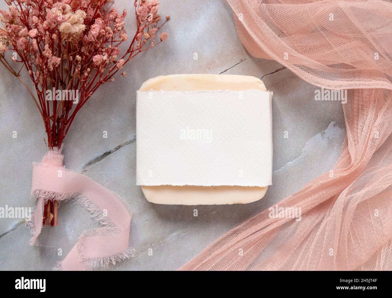 Handmade Soap bar laying on a marble table with paper wrapping package, with pink tulle fabric and dried flowers around, top view. Soap mockup flat la Stock Photo