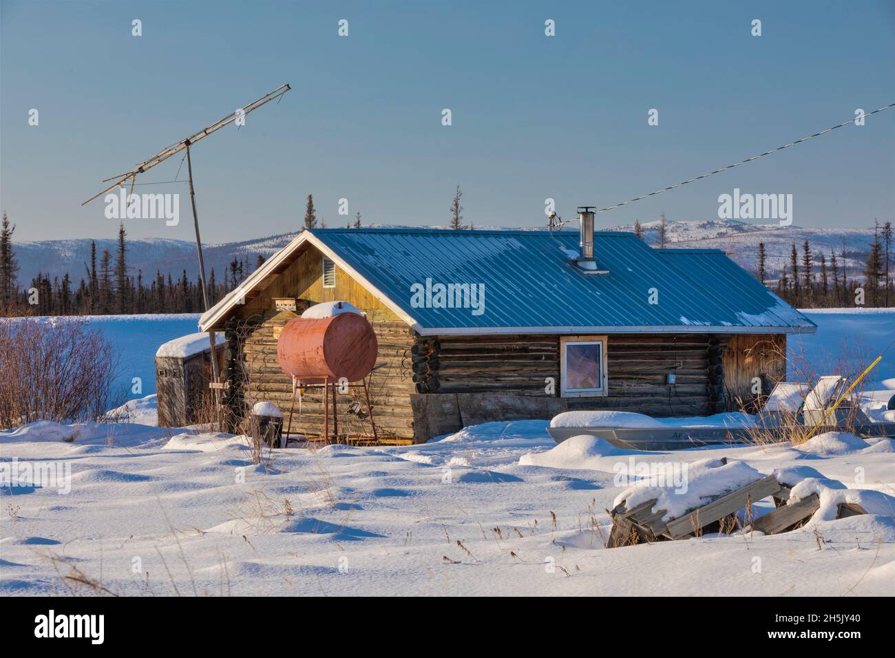 A log cabin is surrounded by snow, TV antenna and heating oil drum in winter, Nulato, Interior Alaska, USA; Nulato, Alaska, United States of America Stock Photo
