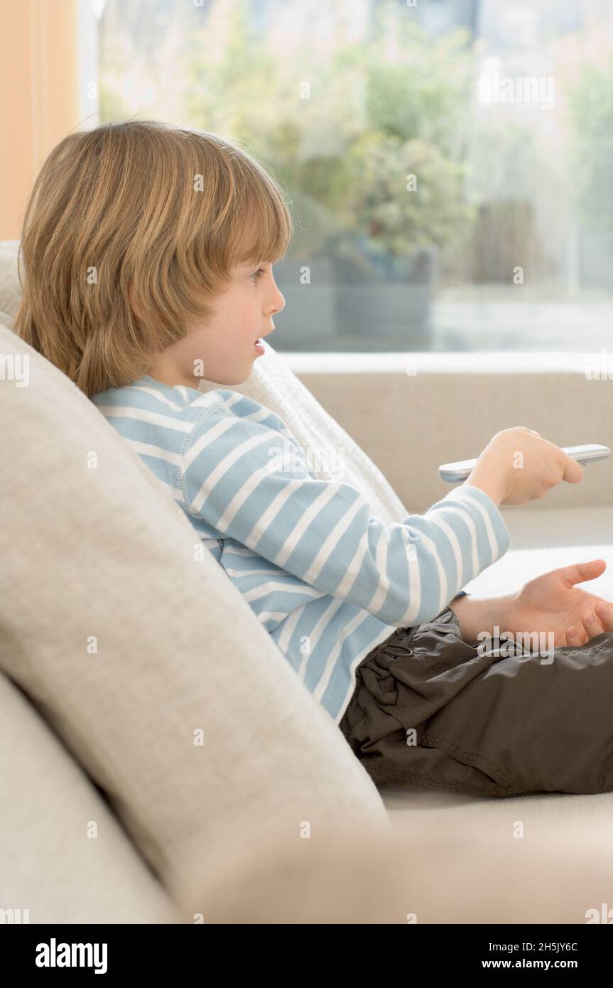 Boy sitting on sofa using remote control at home; Germany Stock Photo