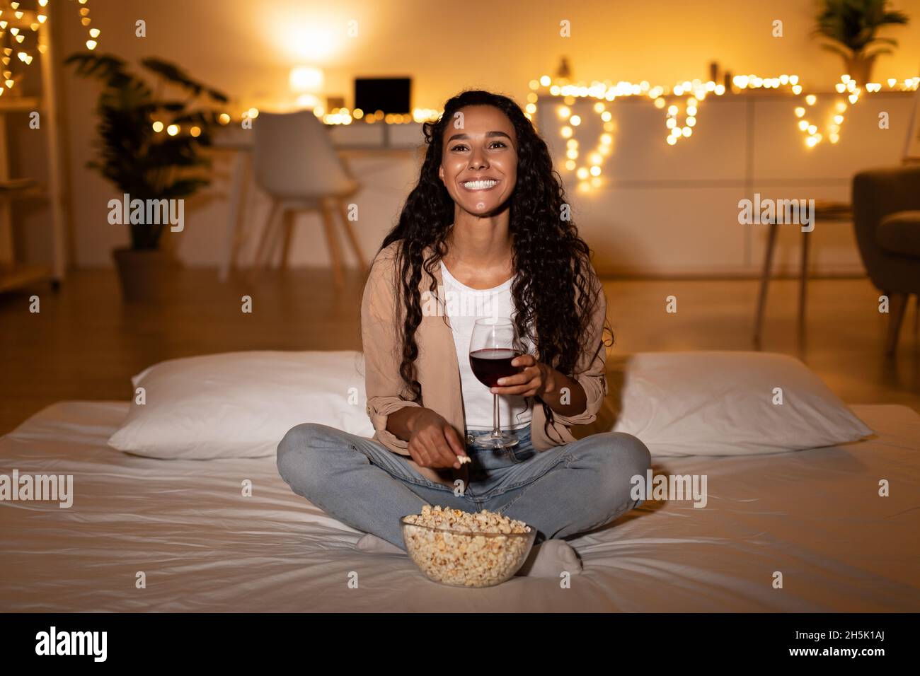 Happy Evening Alone. Joyful Female Drinking Wine And Eating Popcorn Watching Film Sitting On Bed At Home, Smiling To Camera. Cozy Domestic Weekend Lei Stock Photo